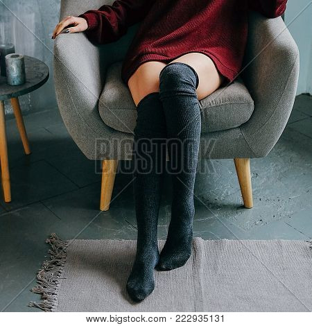 Young adult relaxed woman in casual outfit made of bordeaux sweater and kneesocks sitting in grey armchair in minimalist modern interior