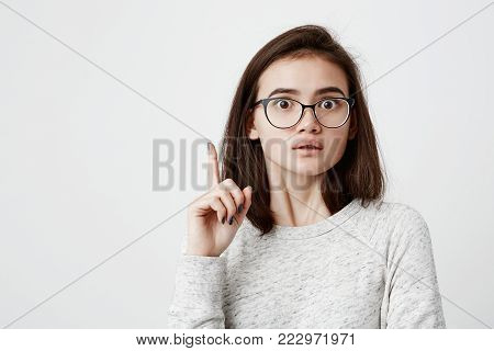 I have a great idea Smart quick-witted pretty girl with dark straight hair wearing eyeglasses keeping finger pointed upwards. Cute female teenager with popped eyes, making gesture with index finger
