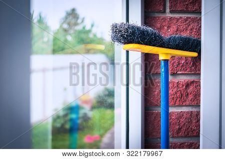Yellow, Blue And Black Poly Push Broom Leaned Against The Glass And Red Brick Wall In The Mud Room. 