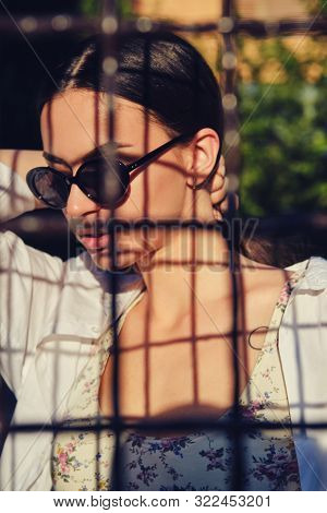 Portrait Of Girl In Sunglasses Posing In City Behind A Trellised Fence. Dressed In Top With Floral P