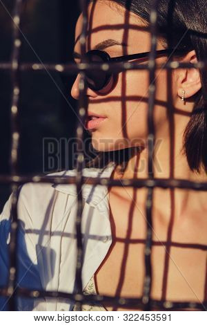 Portrait Of Girl In Sunglasses Posing In City Behind A Trellised Fence. Dressed In Top With Floral P