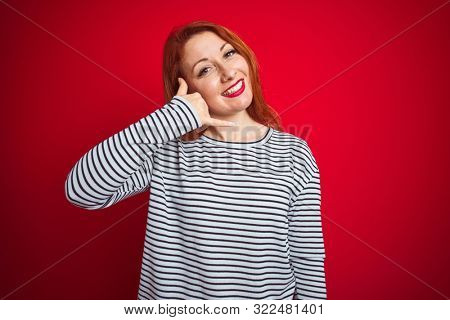 Young redhead woman wearing strapes navy shirt standing over red isolated background smiling doing phone gesture with hand and fingers like talking on the telephone. Communicating concepts.