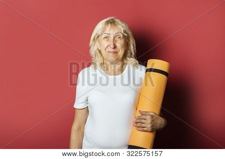 Old Woman Holds A Karemat Or Yoga Mat On A Pink Background. Concept Of Gymnastics, Fitness, Yoga For