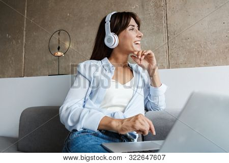 Image of a pleased optimistic young woman indoors at home using laptop computer listening music with headphones sit on sofa in livingroom looking aside.