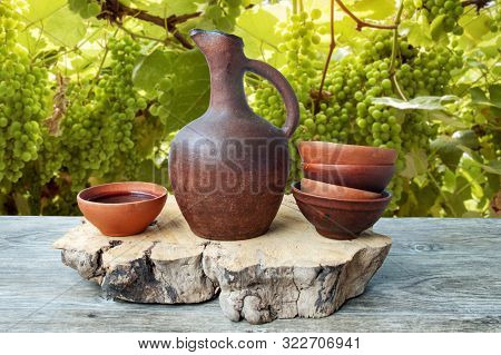 A Traditional Georgian Wine Jug And Clay Cups Filled With A Red Wine On The Wooden Table With Grapes