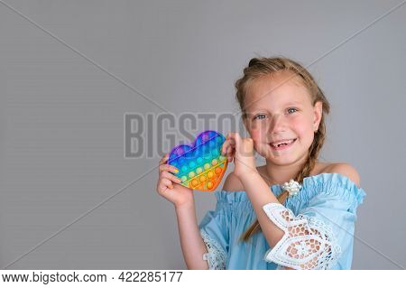 Smiling Child Looking At The Camera And Holding A Rainbow Pop It Fidget Toy Heart Shaped. Push Bubbl