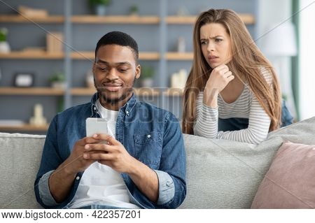 Mistrust. Attentive Lady Looking At Black Boyfriend While He Texting On Smarthone