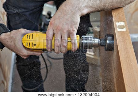 Hands woodworker with yellow electric drill boring large hole of door lock using hole saw drill bits sawdust flying around close-up.