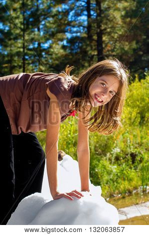 A smiling girl climbs on a block of ice on a sunny day. She is wearing a sleevless shirt and a big smile.