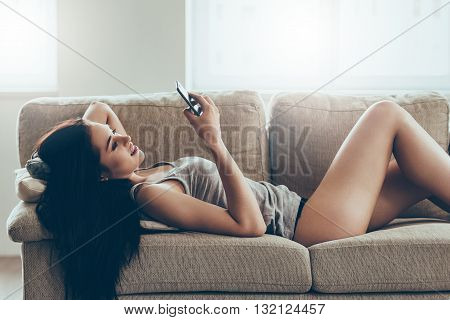 Typing message to him. Beautiful young woman in panties and tank top holding smart phone and looking at it with smile while lying on couch at home
