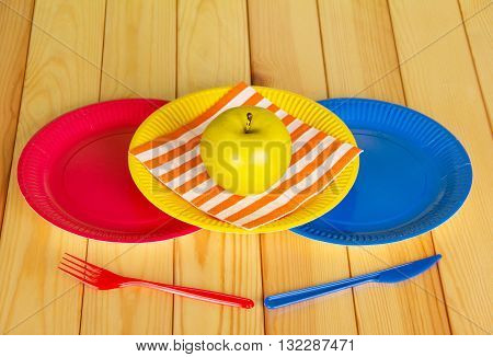 Colored disposable plates, plastic knife, fork, napkin and apple on a background of light wood.