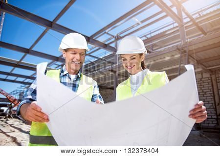 Build together. Young cheerful arhitects outdoors while construction works looking blueprints and building plans with a crane on a background