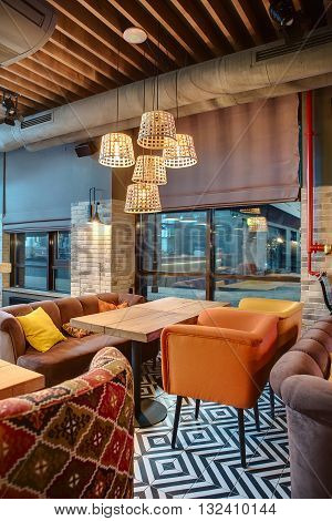 Nice room in a mexican restaurant in a loft style. There are several wooden tables with multi-colored chairs and sofas. On the sofas there are color pillows. Over the tables there are glowing wicker lampshades. On the back wall there are windows 