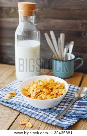 Tasty corn flakes in white bowl with bottle of milk. Rustic wooden background with plaid blue napkin. Healthy crispy breakfast snack.