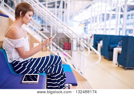 Young caucasian female designer with short brwon hair sitting on a colourful cushion in a co working space while messaging on her smartphone with her tablet by her side.