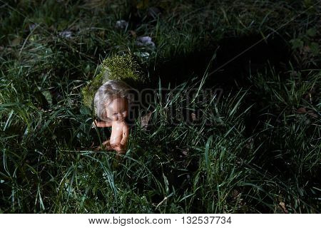 Scary doll lying at dark forest. Conceptual scene of child abuse. Crime scene.