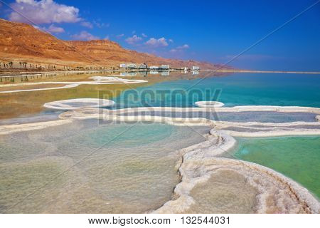 The path from salt picturesquely curls in salty water. Hotels are reflected in smooth water ashore. Israeli coast of the Dead Sea