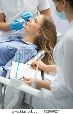 Gorgeous girl with opened mouth in blue shirt. Next to her there is a dentist in a white uniform with blue gloves and a female assistant in a white uniform with blue mask. Assistant writes on the paper in the paper holder. Dentist explores girl's teeth wi