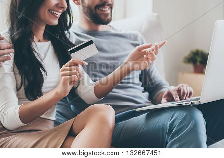I want this! Close-up of beautiful young woman holding credit card and pointing laptop with smile while sitting together with her husband on the couch