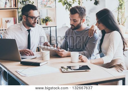 And your signature here. Confident young man signing some document while sitting together with his wife and man in shirt and tie