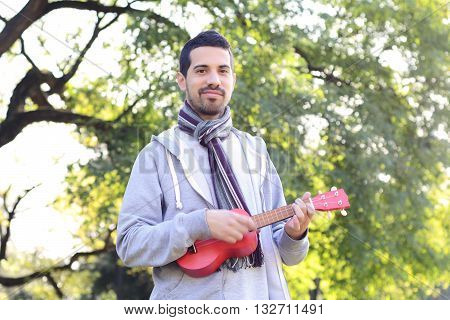 Portrait of young handsome man playing the ukelele in a park. Outdoors.