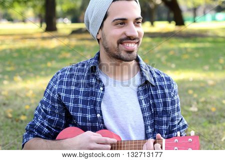 Portrait of young handsome man playing the ukelele in a park. Outdoors.