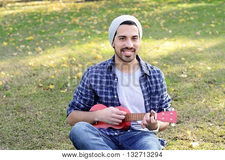 Portrait of young handsome man playing the ukelele in a park. Outdoors.