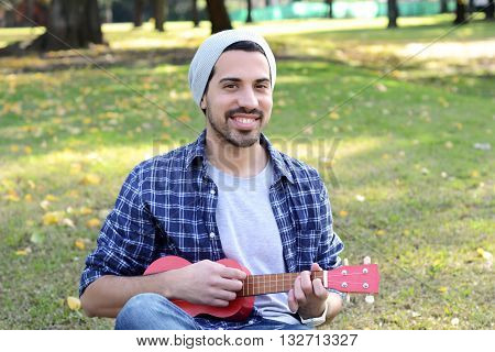 Portrait of young handsome man playing the ukelele in a park. Outdoors.