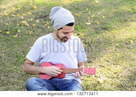 Portrait of young handsome man playing the ukelele in a park. Outdoors.