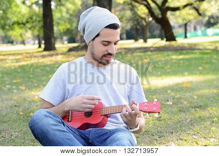 Portrait of young handsome man playing the ukelele in a park. Outdoors.