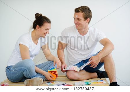 Young people sit on wooden pallet against a white wall and choose color for coloring of the room. The guy looks at the girl and smiles. She examines options. On her face a smile.
