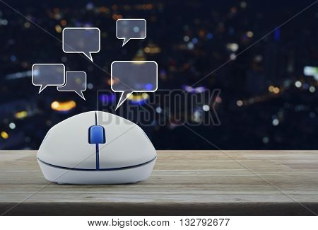 Wireless computer mouse with social chat sign and speech bubbles on wooden table in front of blurred light city tower Social network concept