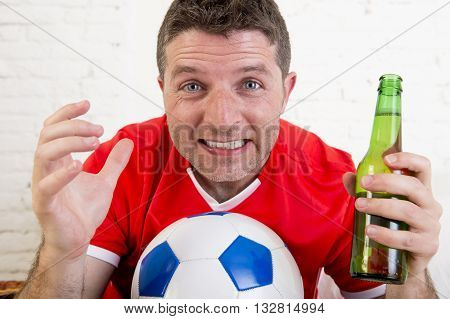 close up face of young fan man watching football game on television wearing team jersey suffering nervous and stress on sofa couch at home holding soccer ball and with beer bottle