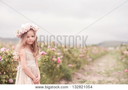 Smiling kid girl 4-5 year old posing in rose garden wearing stylish dress and flower hairband outdoors. Looking at camera. Childhood.