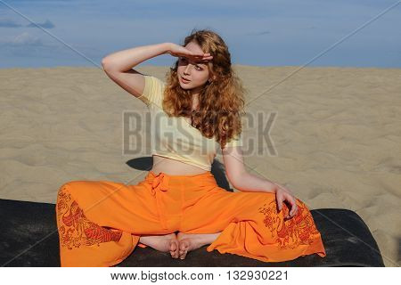 Young slim redhead woman sitting in purna titli yoga pose on the beach. butterfly pose. on the background of the sand and sky