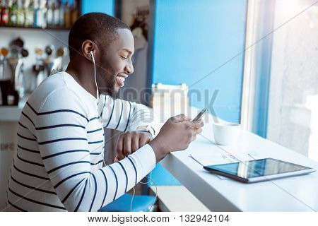 Musical guy.  Smiling and happy young man using his mobile phone and headphones to listen to music while being in a bar and drinking coffee