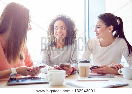 Gossiping together. Cheerful and delighted young women sitting and discussing different things while being in a cafe and using their cell phones