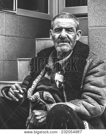 Homelessness Concept. Old Poor And Homeless Gypsy Man Sitting On The Stairs On The Street Smoking Ci
