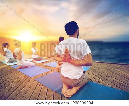 fitness and healthy lifestyle concept - group of people doing yoga reverse prayer pose on wooden pier over sea background