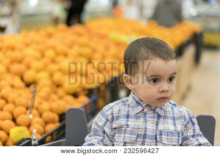 A Cute Boy Dressed In A Plaid Shirt In A Supermarket In A Trolley. Against The Background Of Appelle