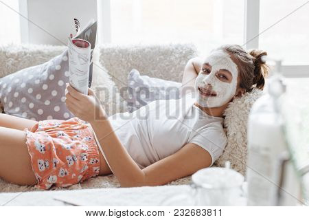 12 years old preteen relaxing on the sofa and making clay facial mask. Teenage girl doing anti blemish skin treatment. Morning skin care routine.