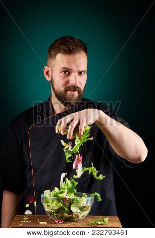 Portrait Of A Handsome Mischevious Male Chef Cook In Black Uniform Making Delicious Vegetable Salad 