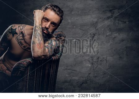 Portrait Of Bearded Male With Tattooed Naked Torso Sits On A Chair Over Dark Grey Background.