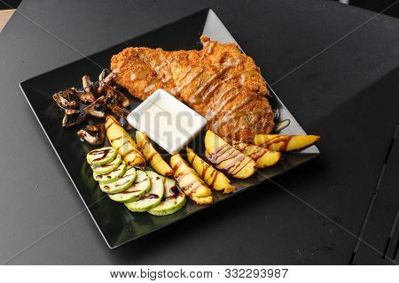 Large Schnitzel With Baked Golden Potatoes, Mushrooms And Zucchini On A Black Background. Top View. 