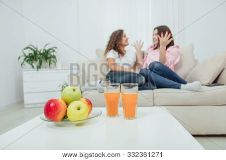 Pile Of Fresh Yummy Apples And Two Glasses Of Juice On The Table On The Forefront. Two Amazing Girls