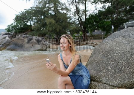 Young Woman Sitting On Sand Near Sea And Stones, Chatting By Smartphone.