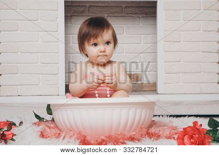 A Little Girl Is Bathing In A Bath With Flowers By The Fireplace. A Child Washes In A Basin Among Re