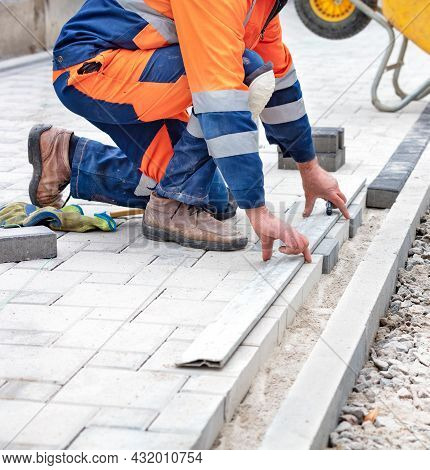A Worker In Blue And Orange Overalls Measures Out A Piece Of Paving Slabs With A Black Marker And An