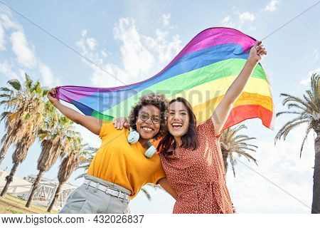 Young Lesbian Couple Celebrates Pride Day Looking At Camera With Rainbow Flag. Concept Of Homosexual