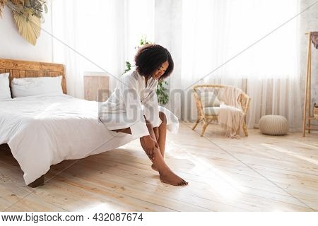 Beautiful Afro Woman In Dressing Robe Touching Her Leg With Soft Skin After Depilation, Sitting On B